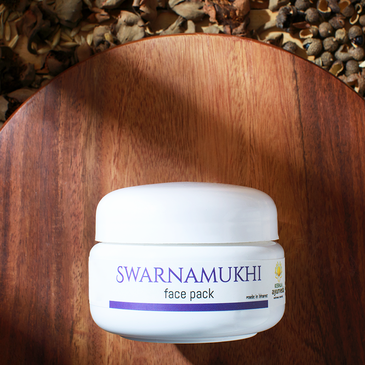 Close-up top view of a white jar of Kerala Ayurveda Swarnamukhi Face Pack on a wooden platter, surrounded by dried herbs. – Kerala Ayurveda India